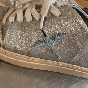 Axel arigatos - Snygga sneakers i silverglittrigt material med ljusgrå mockadetaljer och broderad fågel på sidan. Skorna har vita snören, svart hälparti och platt sula. Perfekta för dig som vill sticka ut med en unik detalj. Box och påse ingår