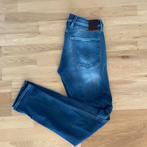 Blå Replay jeans med slitningar - Säljer ett par blå Replay jeans med snygga slitningar och klassisk femficksdesign. Jeansen har normal passform och är tillverkade i denim med lätt stretch. Märkespatch bak i midjan och diskreta detaljer på fickorna.