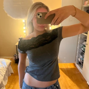 Grå offshoulder topp med spets - Säljer en grå offshoulder topp med svart spetsdetalj över bysten och axlarna. Toppens passform är tight och den är croppad. Perfekt för dig som vill ha en stilren men ändå lite extra look.