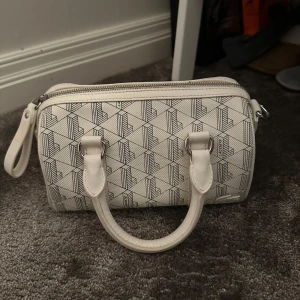 Vit handväska från Lacoste - Säljer en vit handväska från Lacoste. Insidan har ett svart foder och en innerficka med dragkedja. Väskan är köpt utspelande därför €. Väskan är äkta och är köpt från Lacost för ungefär 950kr❤️ 
