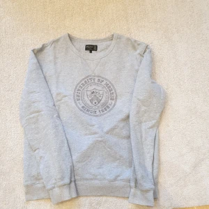 Grå sweatshirt från Morris - Säljer en ljusgrå sweatshirt från Morris med rund hals och tryck på bröstet där det står 'University of Morris Since 1968'. Tröjan har långa ärmar och ribbade muddar vid ärmslut och nederkant. Perfekt för en avslappnad stil.