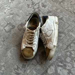 Vita sneakers från Philippe Model   - Säljer ett par vita sneakers från Philippe Model med blå häl och bruna mockadetaljer vid tån och hälen. Skorna har snörning och en platt sula med märkets logga undertill. Perfekta för en avslappnad stil.