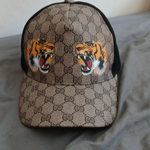 Gucci keps med tigertryck - Säljer en snygg keps från Gucci med klassiskt GG-mönster och två tigrar framtill. Kepsen har svart mesh baktill och justerbart band med gröna och röda ränder. Perfekt accessoar för att sticka ut. Pris kan också diskuteras 