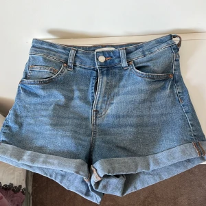Blå jeansshorts från H&M - Klassiska blå jeansshorts från H&M med hög midja och uppvikta benslut. Shortsen har fem fickor och stängs med knapp och dragkedja. Perfekta för varma dagar och enkel att matcha med olika toppar.