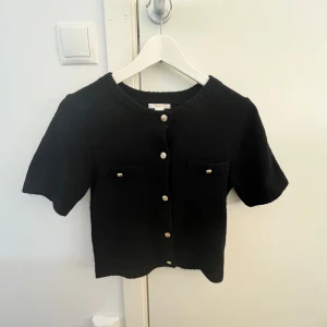 Svart stickad kofta från Gina Tricot - Säljer en svart stickad kofta från Gina Tricot med korta ärmar och dekorativa guldknappar framtill. Koftan har två små fickor på bröstet och ribbade kanter. Perfekt att styla till olika outfits!