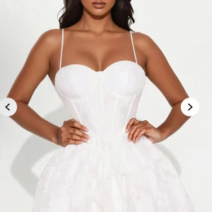 Vit kort klänning med smala axelband - Superfin vit kortklänning med smala axelband och markerad byst. Klänningen har en lätt utsvängd kjol och subtilt blommigt mönster i tyget. Perfekt för sommarens tillfällen!prislappen är på och helt oanvänd 