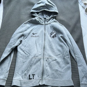 Grå AIK hoodie från Nike - Grå hoodie med dragkedja från Nike, AIK-logga på bröstet och Stadium-tryck. Har huva och fickor framtill samt tryck med 'LT' på ena sidan. Baksidan har ett stort 'hitta.se'-tryck. Perfekt för dig som gillar fotboll och AIK.