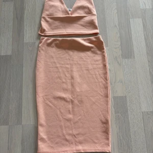 Persikofärgad tvådelad klänning från NLY One - Säljer en persikofärgad tvådelad klänning från NLY One i crepe-material. Toppen är ärmlös med djup v-ringning och kjolen är midilång med tight passform. Perfekt för dig som vill sticka ut med en stilren look. Strl XS, passar även S. Använd en gång 