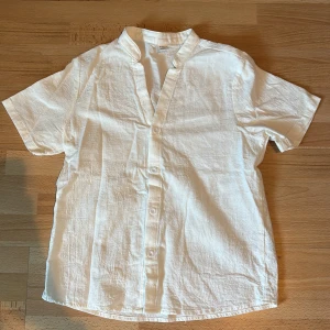 Vit linneskjorta  - Säljer en stilren vit skjorta i linne med korta ärmar och ståkrage. Skjortan har knäppning framtill med vita knappar och en avslappnad passform. Perfekt för varma dagar och enkel att matcha med olika outfits.
