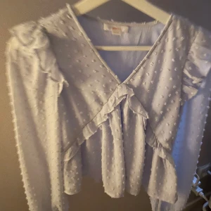 Vit blus med volang och prickigt mönster från Gina Tricot - Säljer en söt vit blus från Gina Tricot med volangdetaljer och diskret prickigt mönster i tyget. Blusen har långa ärmar och v-ringad hals. Perfekt för en trendig och feminin look.