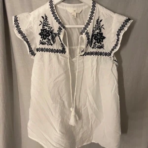 Vit blus med broderade detaljer - Säljer en vit blus med svart/blå broderade blommor vid ärmar och hals. Blusen har korta ärmar och knytband framtill med tofsar. Perfekt för en bohemisk stil. Storlek S