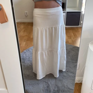 Vit långkjol från Kappahl - Säljer en vit långkjol från Kappahl med smock i midjan och lätt rynkad kjol. Kjolen är luftig och har en enkel, stilren design som passar perfekt till varma dagar.