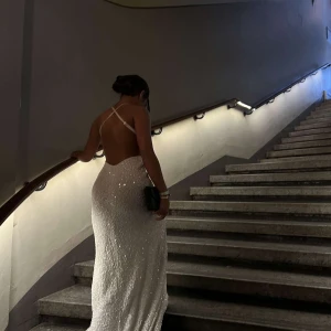 Vit glittrig långklänning med öppen rygg - Säljer en elegant vit långklänning med glittrigt tyg och öppen rygg. Klänningen har smala axelband som korsas i ryggen och en v-ringad front. Perfekt för speciella tillfällen när du vill sticka ut. Även insydd vid låren går att sprätta upp om man vill. Köpt från dressroom.