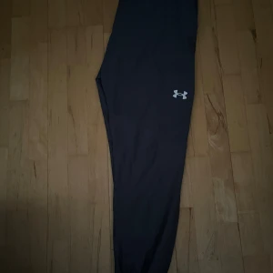 Mörkgråa  träningsbyxor från Under Armour - Mörkgråa byxor från Under Armour med logga på låret. Perfekta för träning med tight passform och stretchigt material som ger bra rörelsefrihet. Storlek XL