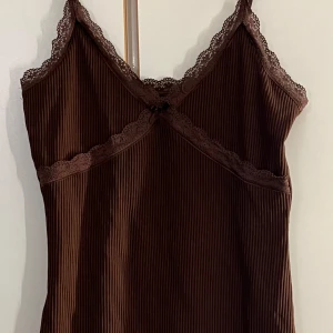 Brun ribbad linnetopp med spets - Säljer en brun ribbad linnetopp med tunna axelband och spetsdetaljer längs halsringningen. Toppen har en liten rosett framtill och är tillverkad i ett mjukt, stretchigt material. Perfekt för lager-på-lager eller som den är.
