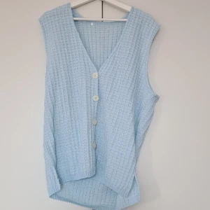Ljusblå stickad väst med knappar - Säljer en ljusblå stickad väst med v-ringning och tre stora knappar framtill. Västen har ett diskret ribbat mönster och är perfekt att styla över en skjorta eller t-shirt. Enkel och stilren design som passar till många outfits.