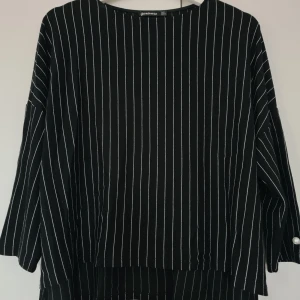 Svart randig blus från Stradivarius - Säljer en svart blus från Stradivarius med vita vertikala ränder. Blusen har trekvartsärmar med dekorativa pärldetaljer vid ärmsluten. Perfekt för en stilren och trendig look.