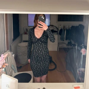 Svart blommig långärmad klänning - Säljer en svart klänning med små vita blommor och djup v-ringning. Klänningen är figurnära och har långa ärmar. Perfekt för dig som gillar en feminin och trendig stil.