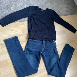 Mörkblå stickad tröja från Polo Ralph Lauren - Säljer en mörkblå stickad tröja från Polo Ralph Lauren med klassiskt kabelstickat mönster och rund halsringning. Tröjan har långa ärmar och en liten röd logga broderad på bröstet. Perfekt för en stilren och tidlös look.