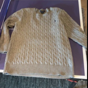 Grå  kabelstickad tröja från Polo Ralph Lauren - Säljer en klassisk grå  kabelstickad tröja från Polo Ralph Lauren. Tröjan har rund halsringning, långa ärmar och ribbade muddar vid ärmslut och nederkant. Perfekt för lager-på-lager och stilrena outfits.