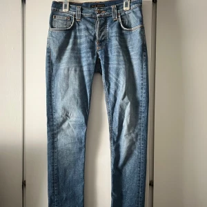 Blå jeans från Nudie Jeans - Säljer ett par klassiska blå jeans från Nudie Jeans med orangea sömmar och fem fickor. Byxorna har normal passform och raka ben, samt dragkedja och knapp i gylfen. Perfekta för dig som gillar stilrena och tidlösa jeans. Har 2 defekter, se bild