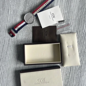 Randig klocka från Daniel Wellington - Säljer en stilren klocka från Daniel Wellington med randigt armband i rött, vitt och blått. Klockan har en silverfärgad urtavla och kommer i originalförpackning med tillhörande dokumentation.