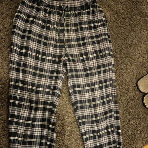 Rutig mjukisbyxa - Säljer ett par bekväma mjukisbyxor med rutigt mönster i svart, grått och vitt. Byxorna har en elastisk midja med snörning för justerbar passform. Perfekta för en avslappnad dag hemma.