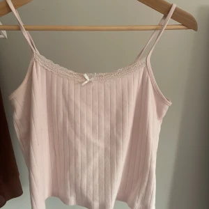 Ljusrosa ribbad linnetopp med spetsdetaljer - Söt ljusrosa linnetopp med smala axelband och ribbad struktur. Toppen har fina spetsdetaljer längs halsringningen och en liten rosett framtill. Perfekt för en romantisk look. Från Gina tricot 