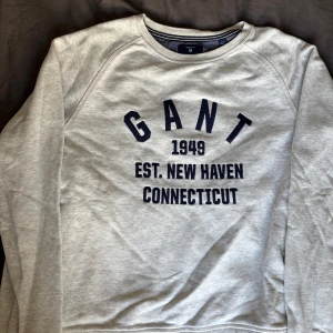 Grå sweatshirt från GANT - Säljer en stilren grå sweatshirt från GANT med tryck i marinblått. Tröjan har rund hals och långa ärmar, perfekt för en avslappnad stil. Passar bra till både jeans och chinos.
