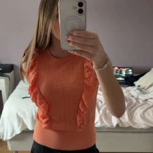 Orange stickad topp med volangdetaljer - Säljer en snygg orange stickad topp med volangdetaljer längs sidorna. Toppen har en rund hals och är ärmlös. Skriv för flera bilder 