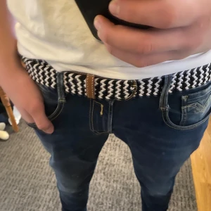 Svartvitt flätat bälte med svarta detaljer - Snyggt flätat bälte i svart och vitt med bruna läderdetaljer och ett silverfärgat spänne. Perfekt för att ge en stilren touch till din outfit. Passar till både jeans och chinos.
