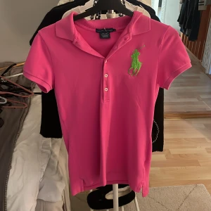 Rosa pikétröja från Ralph Lauren - Säljer en snygg rosa pikétröja från Ralph Lauren med en stor grön logga på bröstet. Tröjan är tillverkad i mjuk pima bomull med en liten andel elastan för extra komfort. Storlek S!