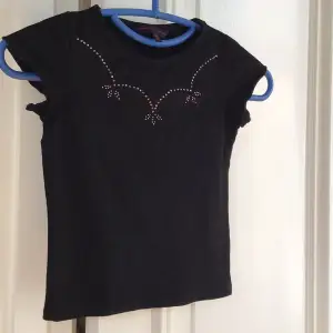 Säljer en svart kortärmad topp från French Connection med dekorativa stenar på framsidan. Perfekt för en stilren look. Passar barn i storlek 6.