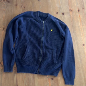 Mörkblå kofta från Lyle & Scott - Säljer en snygg mörkblå kofta från Lyle & Scott med dragkedja och två fickor framtill. Koftan har ett diskret mönster och det klassiska gula logotypmärket på bröstet. Perfekt för kyligare dagar!
