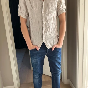 Beige linneskjorta - Säljer en stilren beige skjorta i linne med korta ärmar och knappar framtill. Perfekt för varma dagar med sin lätta och luftiga känsla. Skjortan är i storken S