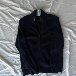 Marinblå kofta från Ralph Lauren - Snygg Marinblå kofta från Ralph Lauren med dragkedja framtill. Den har en ribbad krage och muddar, samt en liten broderad logotyp på bröstet.  Strl 18-20 på ungdom. Hör av er vid frågor! Pris är diskuterbart!