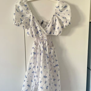 Klänning - Charmig vit klänning med blå blommor och puffärmar. Den har en söt knytning framtill och en luftig passform, perfekt för varma dagar.
