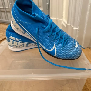 Blå Nike fotbollsskor - Säljer ett par blå Nike fotbollsskor med vit logga och texten 'Just Do It' på sidan. Skorna har snörning och en strömlinjeformad design för optimal prestanda på planen. Storlek 44,5