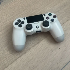 Vit DualShock 4 trådlös handkontroll - Säljer en vit DualShock 4 trådlös handkontroll från Sony. Den har en stilren design med klassiska PlayStation-knappar och en touchpad i mitten. Perfekt för gaming på PlayStation 4. Kontrollen har också inbyggd högtalare och hörlursuttag.