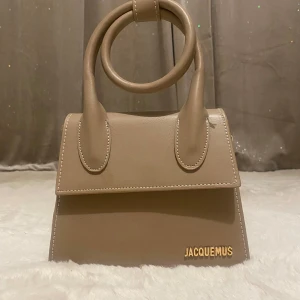 Beige handväska från Jacquemus - Säljer en stilren beige handväska med ett elegant handtag och avtagbar axelrem. Väskan har en minimalistisk design med guldfärgad logotyp på framsidan. Perfekt för att ge din outfit en lyxig touch.