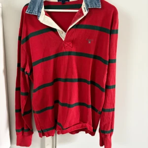 Röd och grön randig pikétröja från GANT - Säljer en klassisk röd pikétröja från GANT med gröna ränder och en stilren krage i denim. Tröjan har långa ärmar och en broderad logotyp på bröstet. Perfekt för en avslappnad och stilren look. Sömmen börjar gå upp lite se bild, annars bra skick 