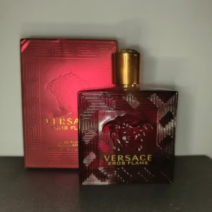 Versace Eros Flame 100ml - Säljer en elegant Versace Eros Flame Eau de Parfum i en djup röd flaska med guldlock. Flaskan har ett stilrent mönster och det ikoniska Versace-emblemet. Perfekt för den som vill ha en doft med karaktär och stil.