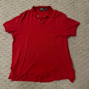 Röd pikétröja från Polo Ralph Lauren - Säljer en klassisk röd pikétröja från Polo Ralph Lauren med broderad logga på bröstet. Tröjan har korta ärmar och en stilren krage med knappar. Perfekt för en avslappnad och stilren look. Storlek XL