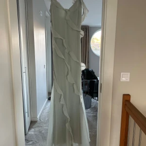 Ljusgrön långklänning från Coast - Elegant ljusgrön långklänning från Coast med volangdetaljer längs hela klänningen. Klänningen har knappar framtill och är perfekt för speciella tillfällen. Den har en luftig och flödande design som ger en romantisk känsla.