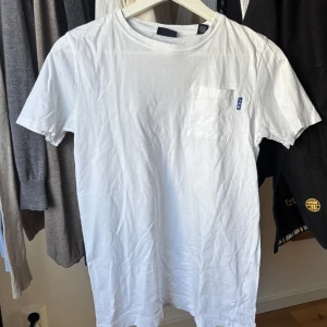 Vit t-shirt från Scotch & Soda - Säljer en stilren vit t-shirt från Scotch & Soda med en liten bröstficka och blå detalj. Perfekt för en avslappnad look.