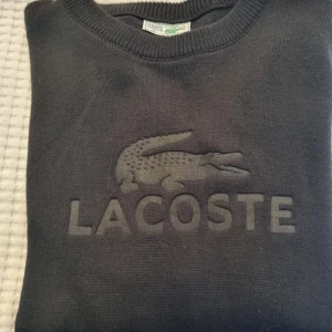 Svart tröja från Lacoste - Svart tröja från Lacoste med det klassiska krokodilemblem broderat på framsidan. Tröjan har långa ärmar och en rund halsringning.