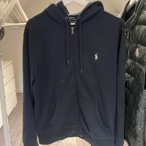 Mörkblå hoodie från Polo Ralph Lauren - Säljer en stilren mörkblå hoodie från Polo Ralph Lauren med dragkedja och justerbar huva. Den har en broderad logga på bröstet och praktiska fickor framtill. Perfekt för en avslappnad look.