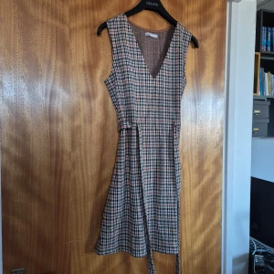 Rutig ärmlös klänning från Cropp - Snygg ärmlös klänning med rutigt mönster i svart, beige och orange. Klänningen har en v-ringad hals och ett knytband i midjan för en smickrande passform. Perfekt för en stilren look!