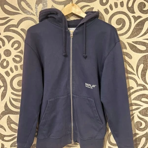 Mörkblå hoodie från Replay - Säljer en snygg mörkblå hoodie från Replay med dragkedja och huva. Den har en klassisk passform och är perfekt för en avslappnad stil. Framtill finns två praktiska fickor och Replay-loggan är diskret placerad på bröstet. Skriv för fler bilder, org pris 2200kr