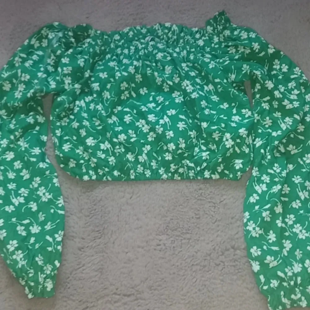 Snygg grön offshoulder blus med vita blommor. Blusen har långa ärmar och en elastisk kant vid axlarna för en bekväm passform. Perfekt för en somrig look!. Yläosat.